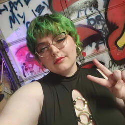 An image of a white person with bright green hair taken from mid-chest up. He is wearing reddish and black winged eyeliner and has large glasses with a rounded bottom and a cat-eye taper on the upper outer corners. He has a septum piercing and two lip pier
